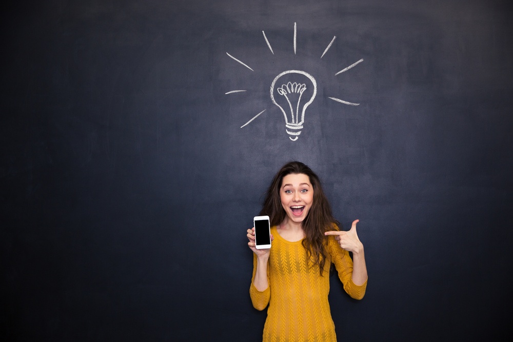 Pretty happy young woman holding and pointing on smartphone blank screen standing over chalkboard background